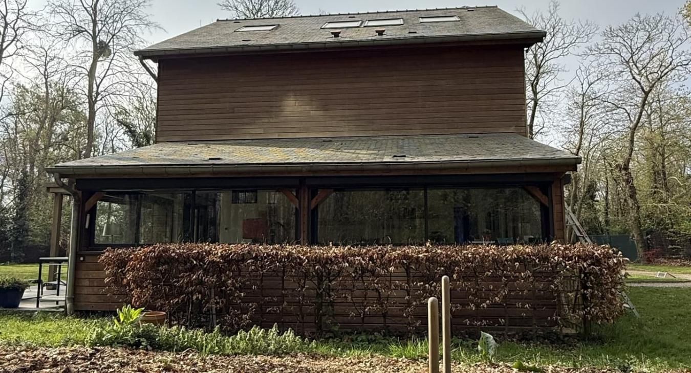 Maison en bois de deux étages avec porche couvert, entourée d'arbres et d'une haie taillée.