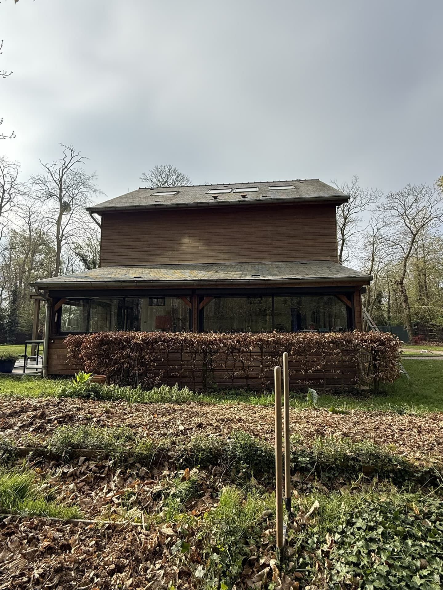 Bâtiment en bois de deux étages avec un rez-de-chaussée vitré, entouré d'arbres et d'une haie. Ciel couvert.