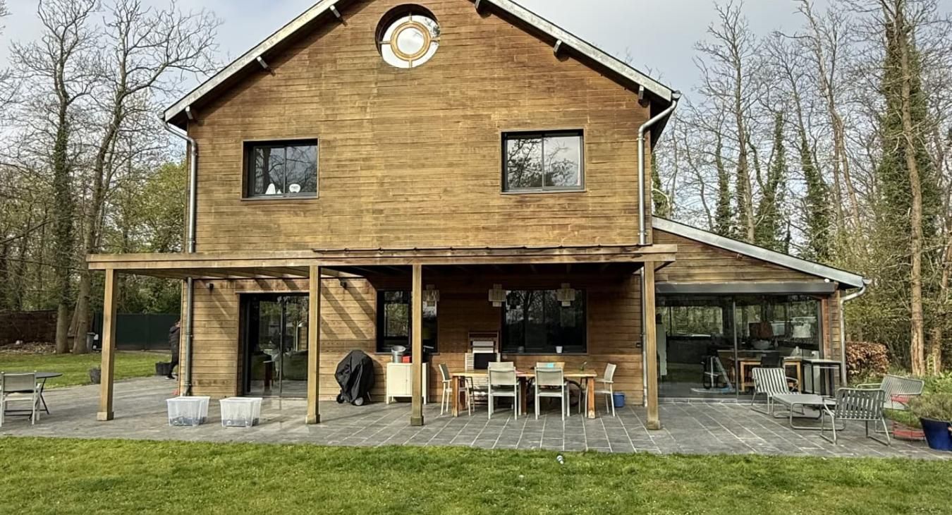 Maison en bois à deux étages avec terrasse, table, chaises et arbres environnants.