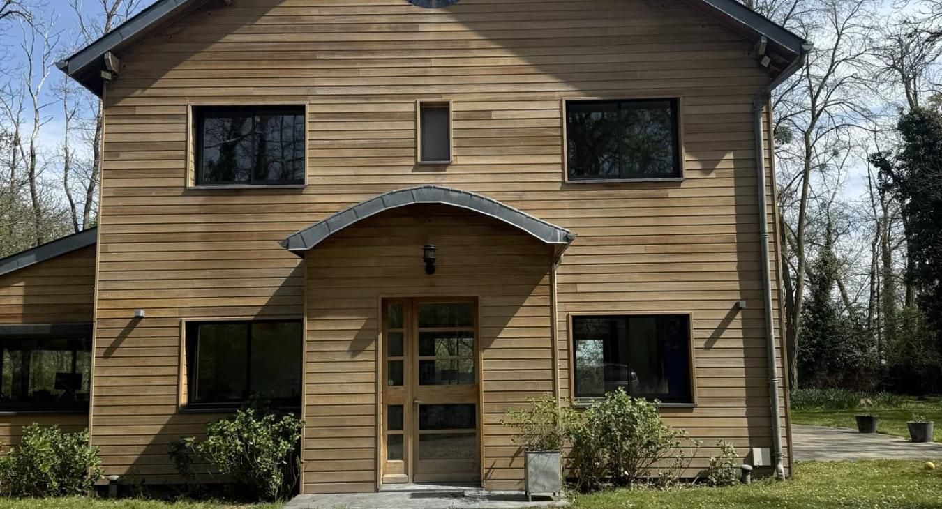 Maison en bois aux fenêtres à cadre noir et auvent d'entrée incurvé, située dans un paysage verdoyant.