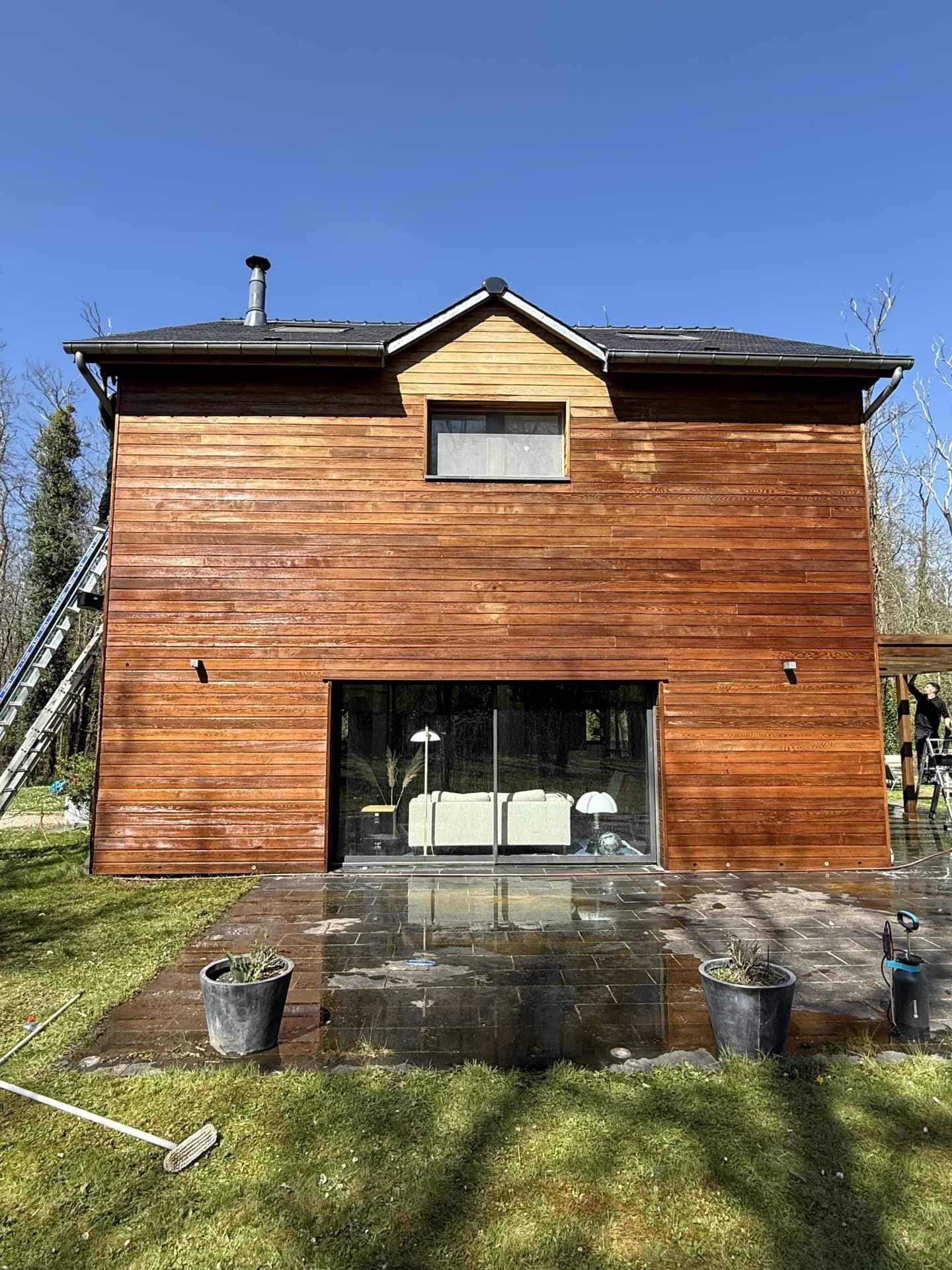 Maison en bois à deux étages avec de grandes portes vitrées et une petite fenêtre, située sur un patio ; journée ensoleillée.