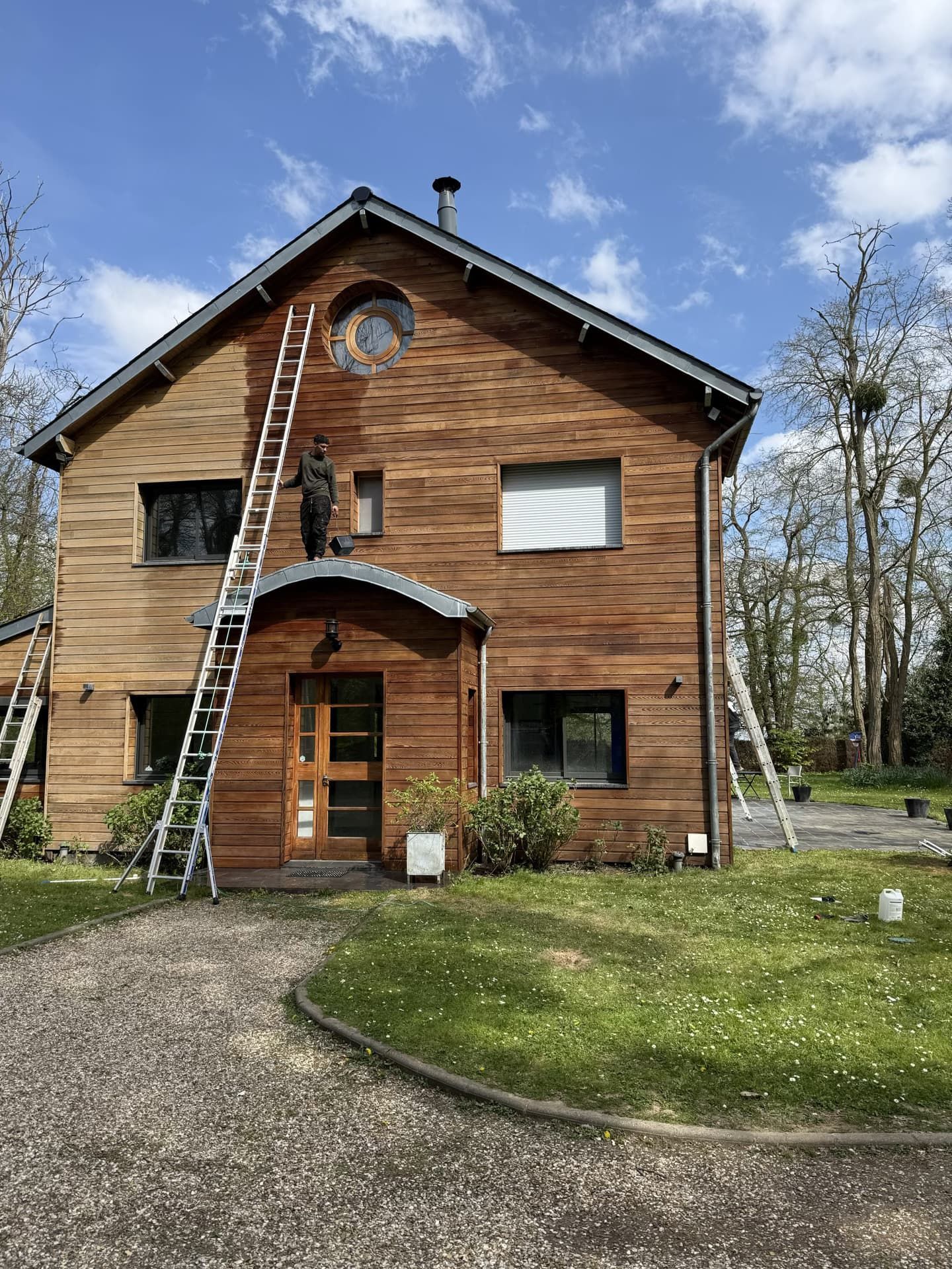 Maison en bois en cours de peinture ; personne sur une échelle. Ciel bleu, arbres et allée de gravier.