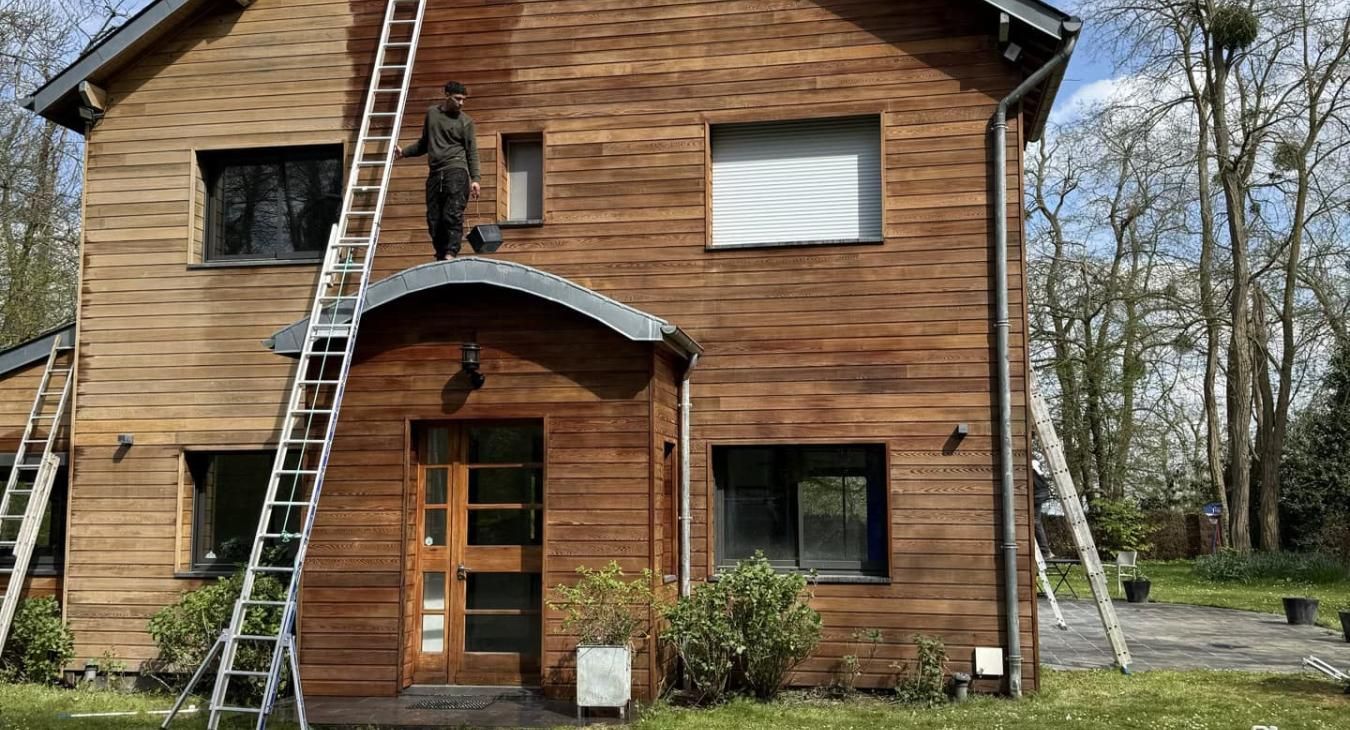 Une personne, perchée sur une échelle, peint une maison en bois à deux étages.