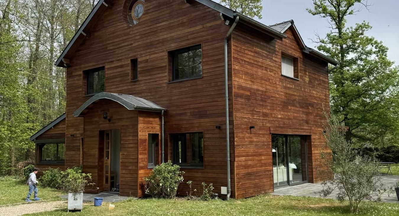Maison en bois de deux étages avec des fenêtres sombres et un petit porche, située dans une cour gazonnée.