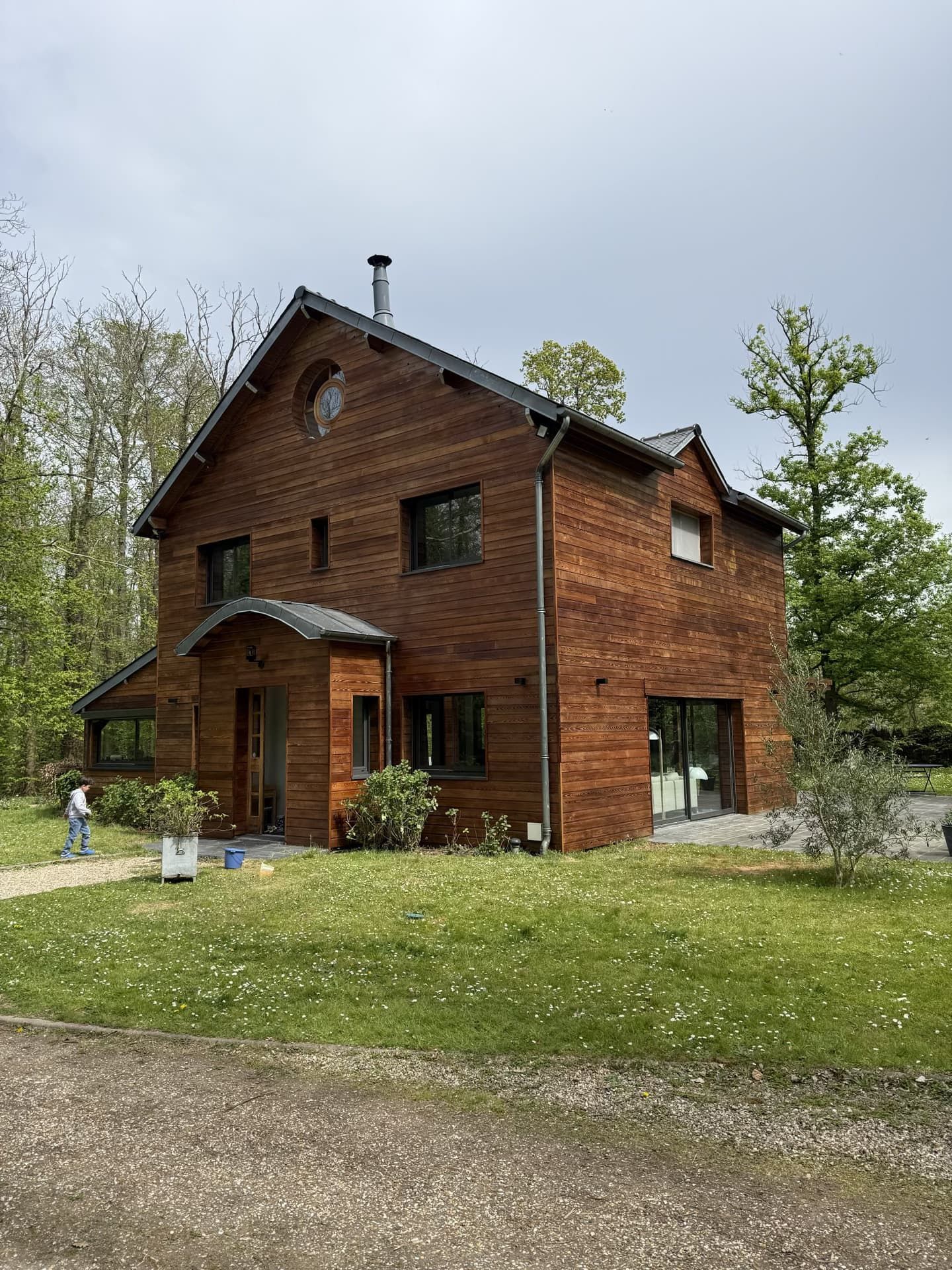Maison en bois à deux étages avec des fenêtres noires, située dans une cour herbeuse entourée d'arbres.