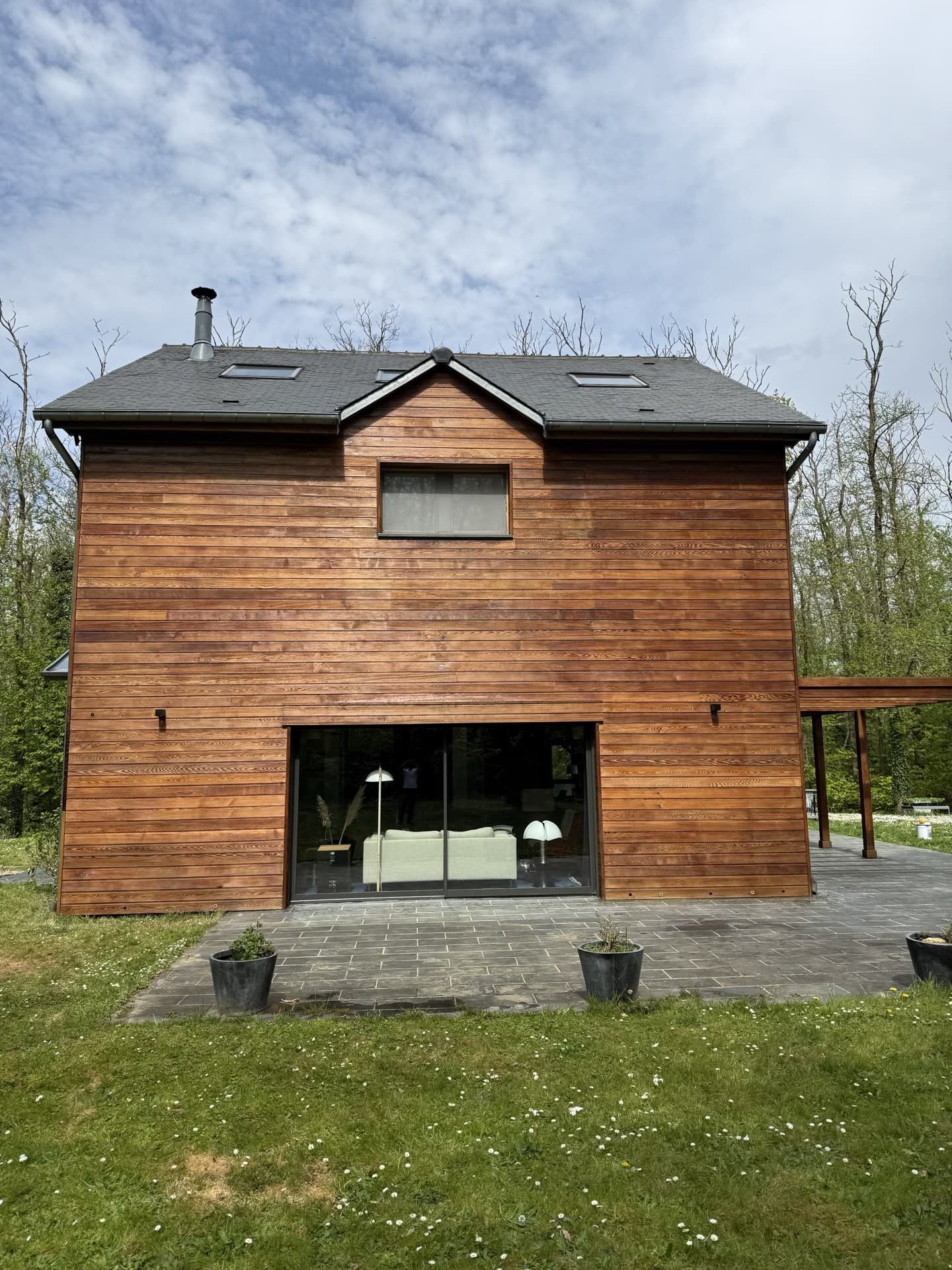 Maison en bois avec de grandes portes vitrées, entourée de pelouse et d'une terrasse en pierre grise.