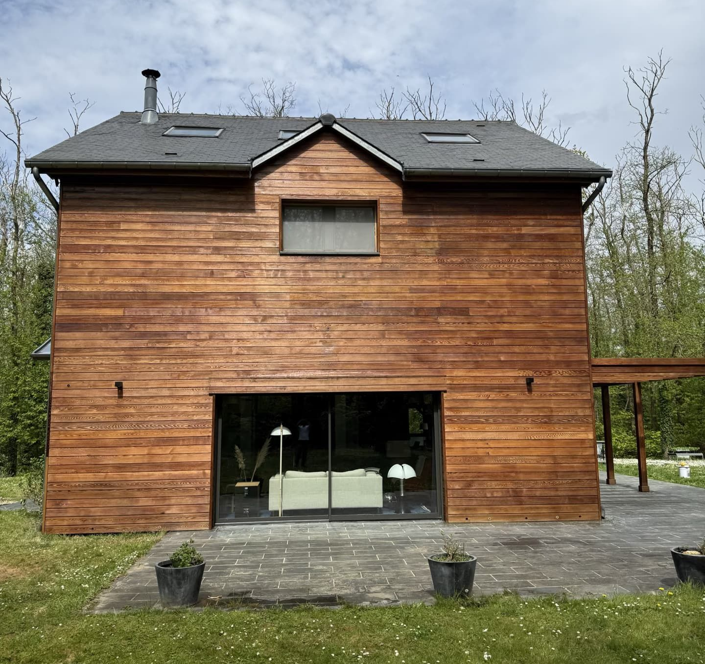 Maison en bois avec une grande façade vitrée, un toit gris et entourée d'arbres.