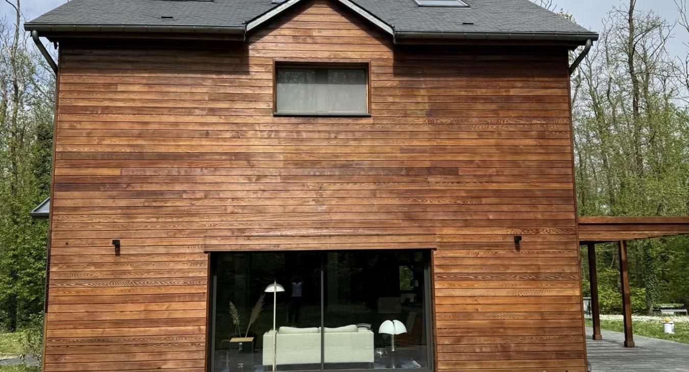 Maison à façade en bois, toit sombre, grande porte vitrée, fenêtre au-dessus de la porte et porche sur la droite.