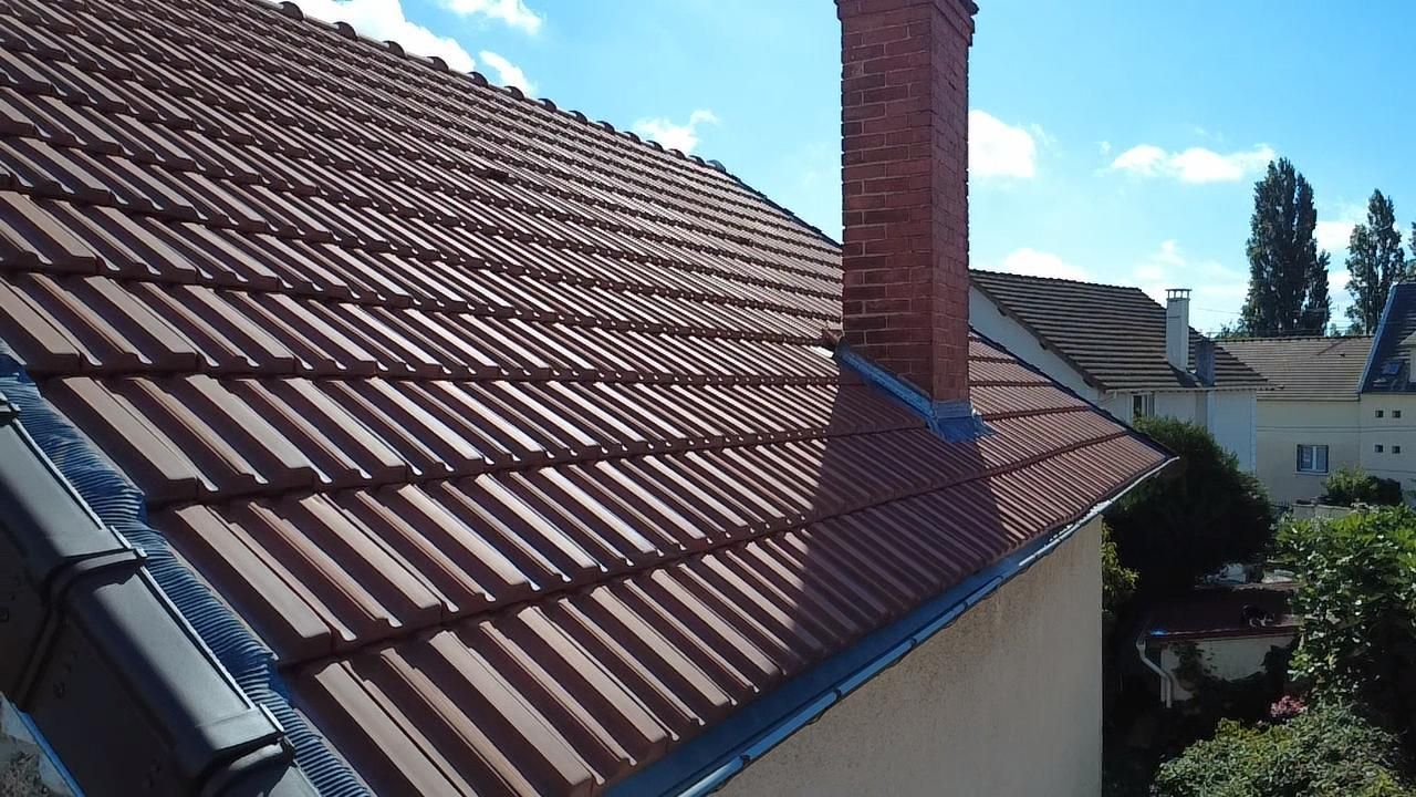 Toit de tuiles rouges avec cheminée en briques se détachant sur un ciel bleu, donnant sur d'autres bâtiments et des arbres.