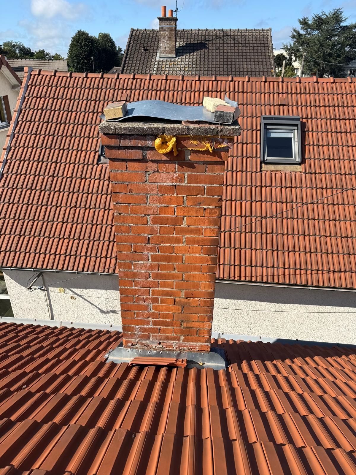 Cheminée en briques sur un toit de tuiles rouges, au sommet endommagé. Lucarne et autre toiture en arrière-plan.