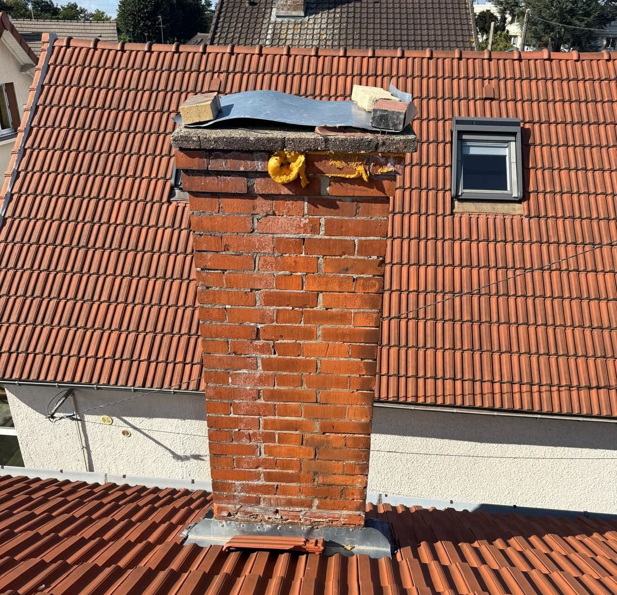 Cheminée en briques sur un toit de tuiles rouges, avec une petite fenêtre et un ciel nuageux visible.