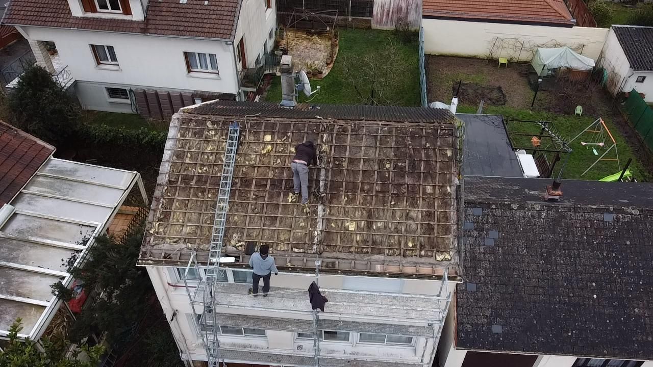 Des ouvriers réparent le toit d'un immeuble ; on aperçoit les échafaudages et les maisons environnantes.