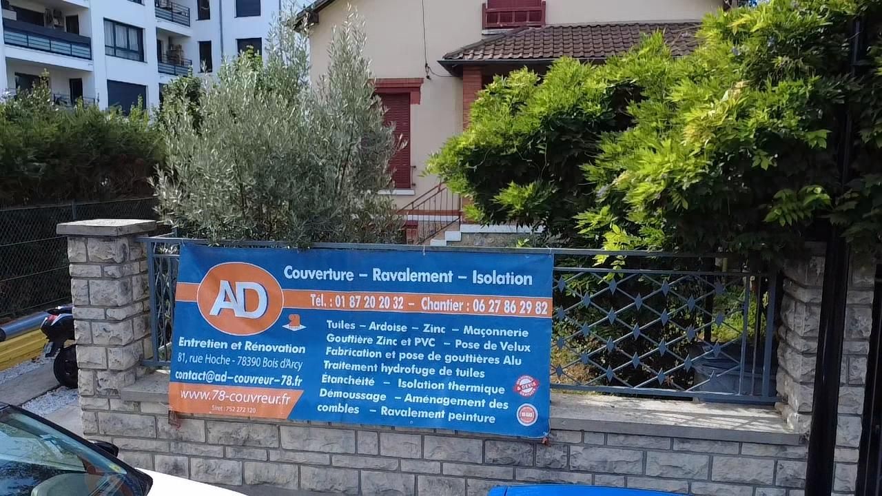 Panneau publicitaire AD Couverture, bleu et orange, devant un bâtiment et des arbres.