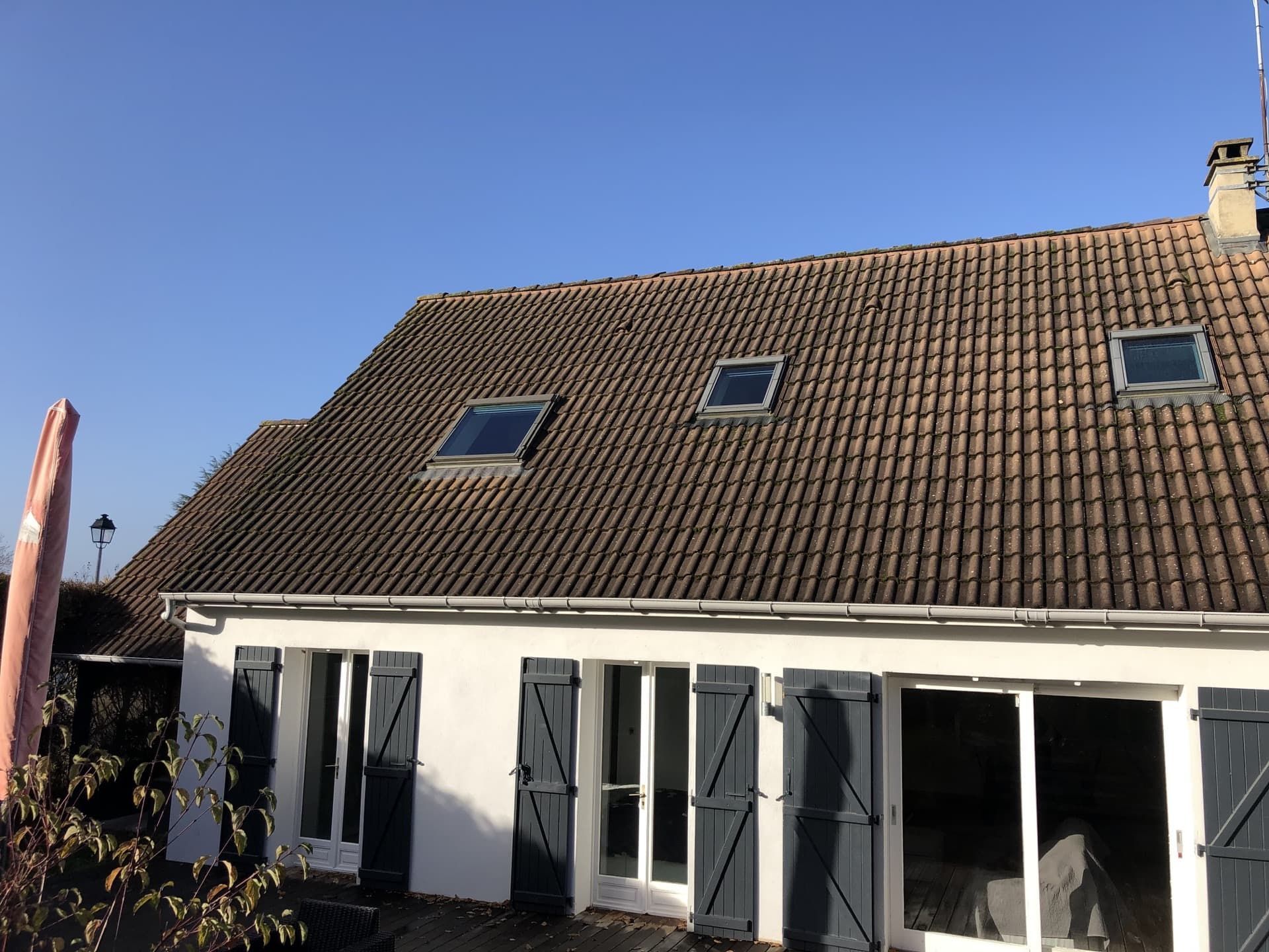 Maison blanche au toit de tuiles brunes, trois puits de lumière, des volets bleus et un ciel d'un bleu limpide.