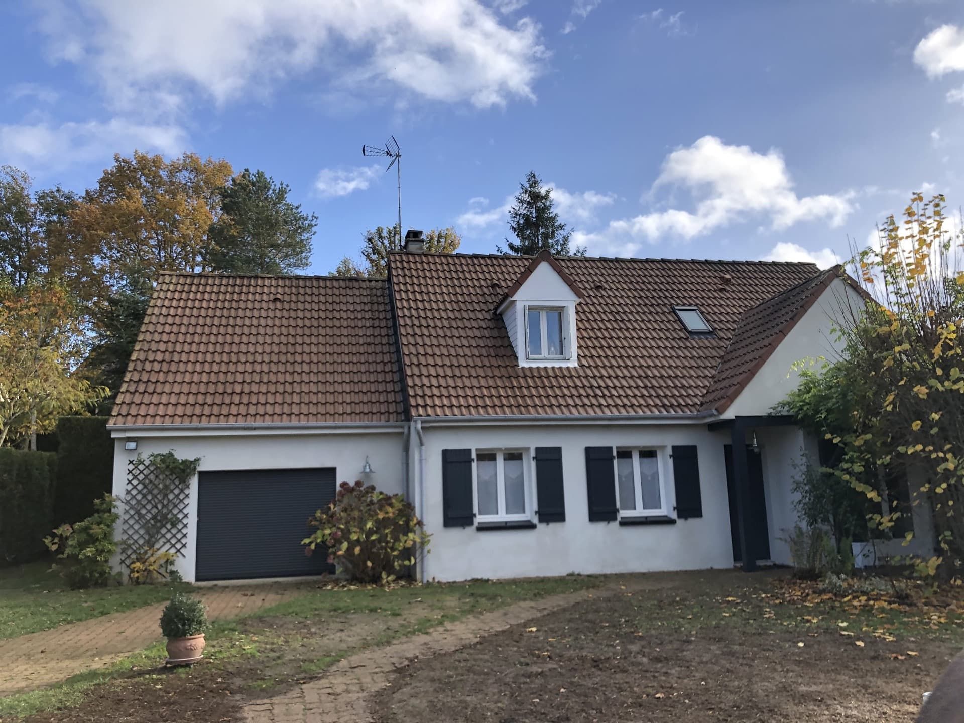 Maison blanche au toit de tuiles brunes, aux volets noirs et à la porte de garage, sous un ciel bleu.