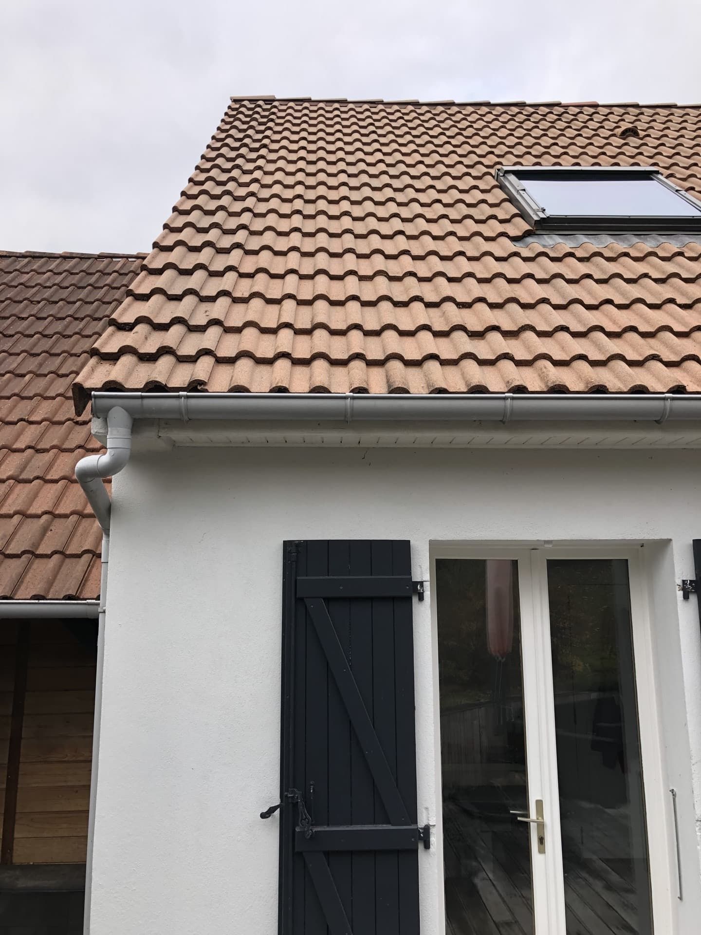 Maison blanche avec toit de tuiles brunes, volets bleu foncé et puits de lumière.