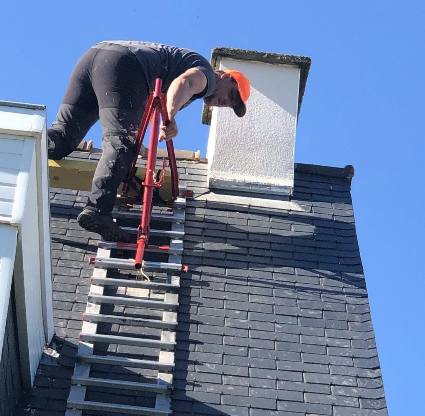 Une personne sur un toit, utilisant une échelle pour travailler près d'une cheminée, sur fond de ciel bleu.