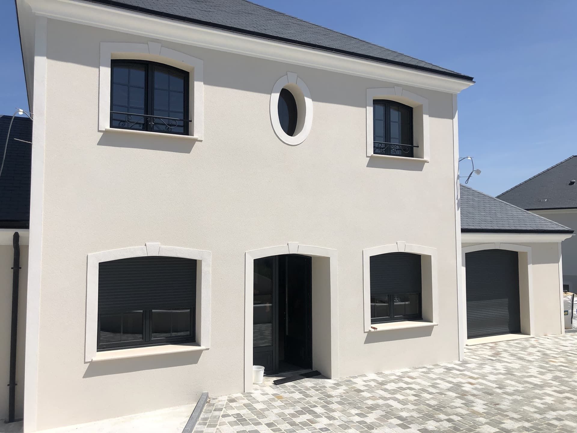 Maison à deux étages avec façade en stuc clair, volets foncés et toit sombre, sous un ciel bleu.