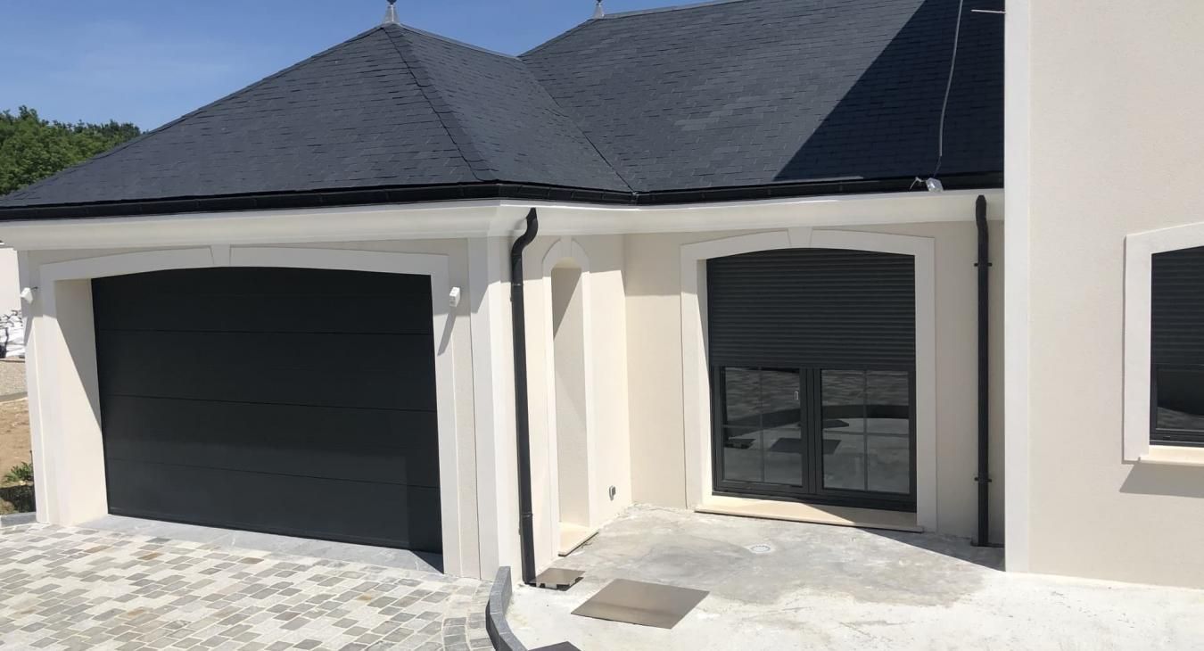 Vue extérieure d'une maison avec une porte de garage gris foncé, des fenêtres aux volets fermés et un toit sombre.