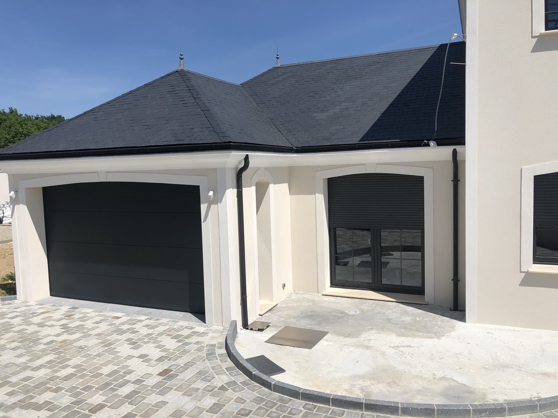Garage avec porte noire, fenêtre cintrée et toit gris foncé sur un extérieur en stuc clair.