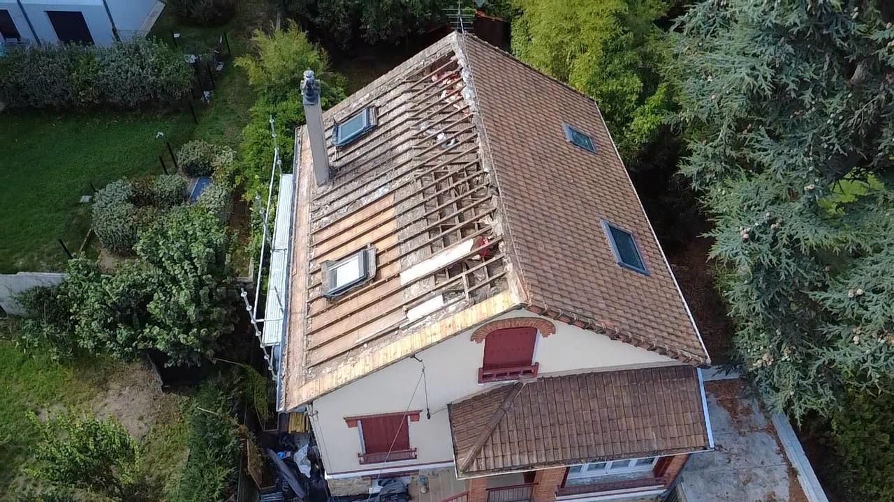 Vue aérienne d'une maison au toit endommagé et aux tuiles manquantes, entourée d'arbres et de verdure.