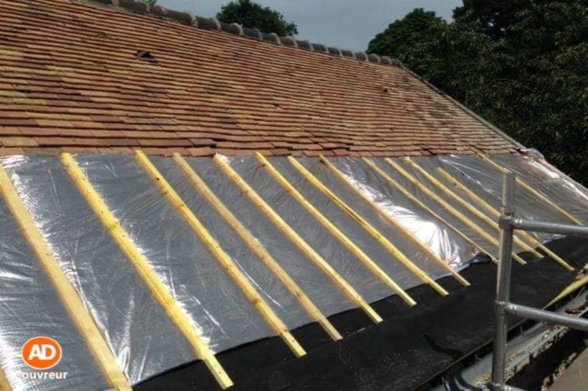 Toiture en construction : tuiles partiellement posées, isolation et chevrons visibles. Échafaudage en équilibre précaire.
