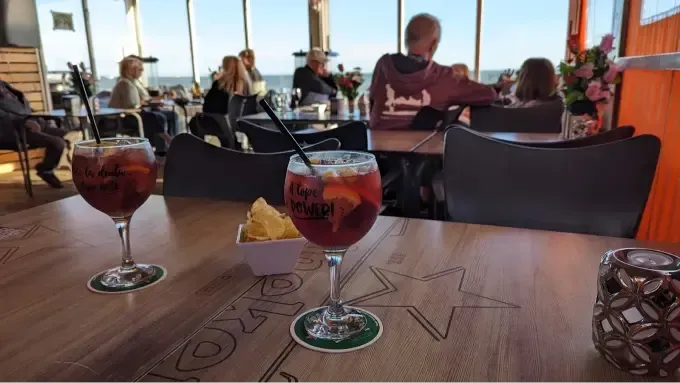 Dos bebidas sobre una mesa de madera en la terraza de un restaurante al atardecer, con comensales