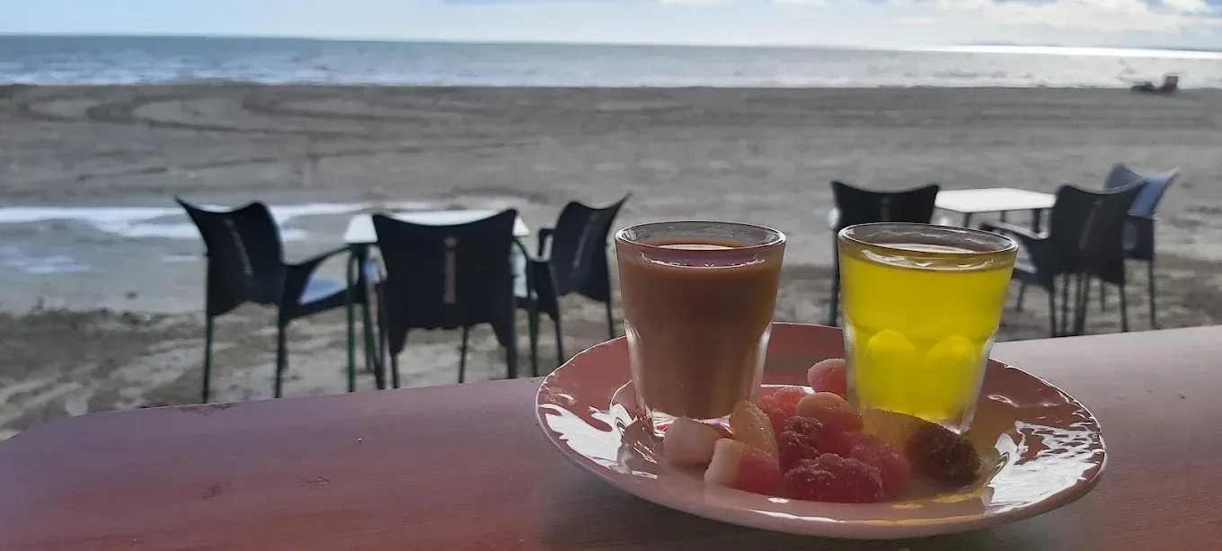 Mesa junto a la playa con dos bebidas y fruta frente al océano y sillas vacías.
