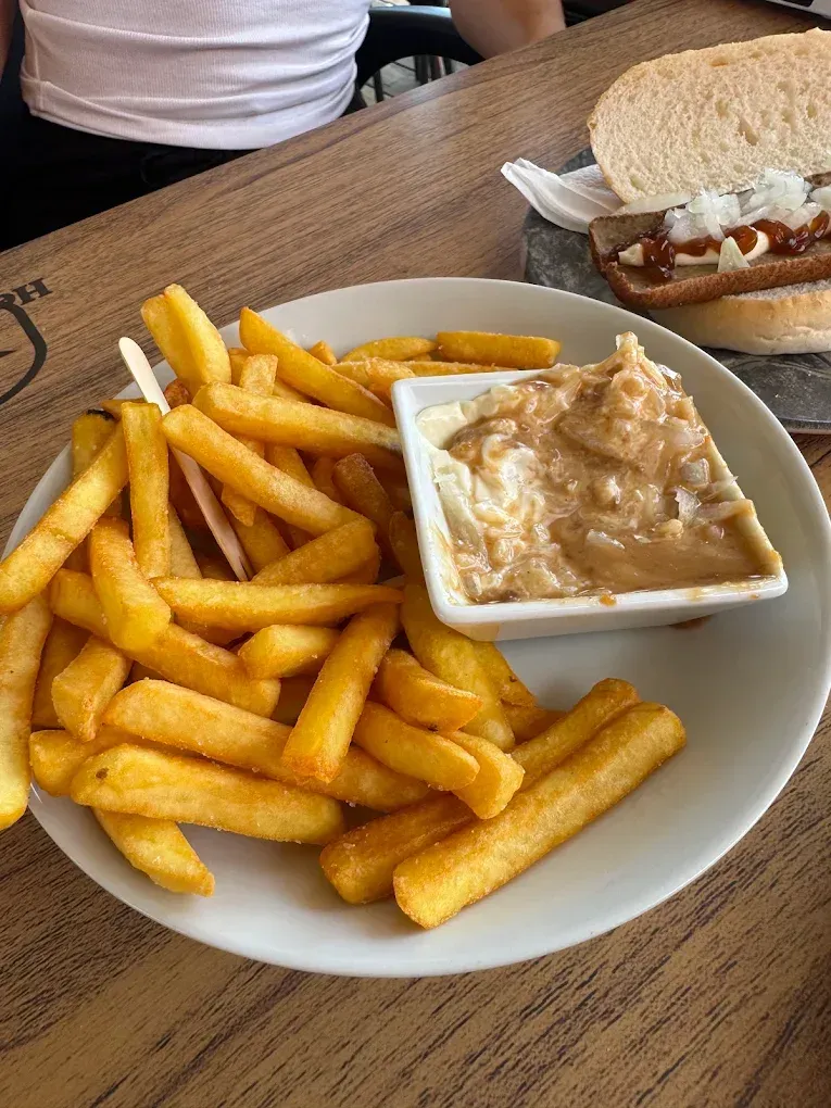 Plato de patatas fritas con un vasito de salsa para mojar sobre una mesa de madera junto a un sándwich.