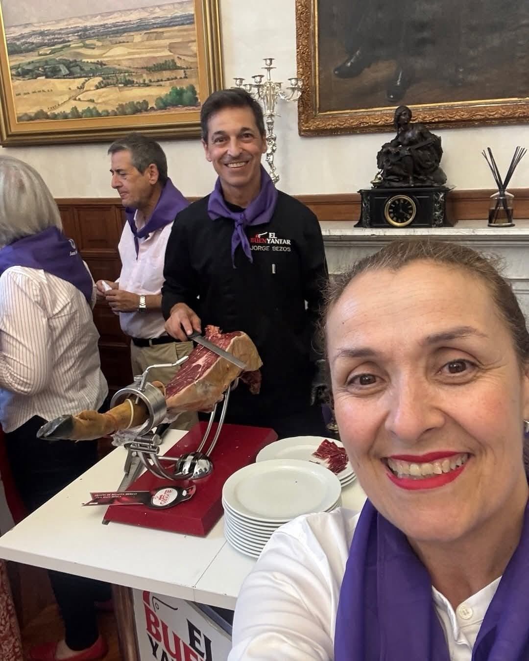 Mujer sonriente tomándose una selfie en una mesa de un evento en interiores con platos, trofeo y pasteles detrás de ella.