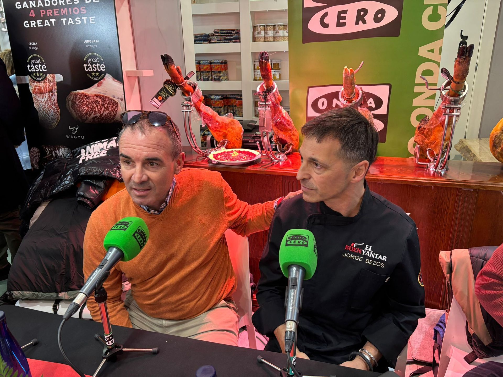 Dos hombres sentados frente a micrófonos en un estudio, uno de ellos con un suéter naranja