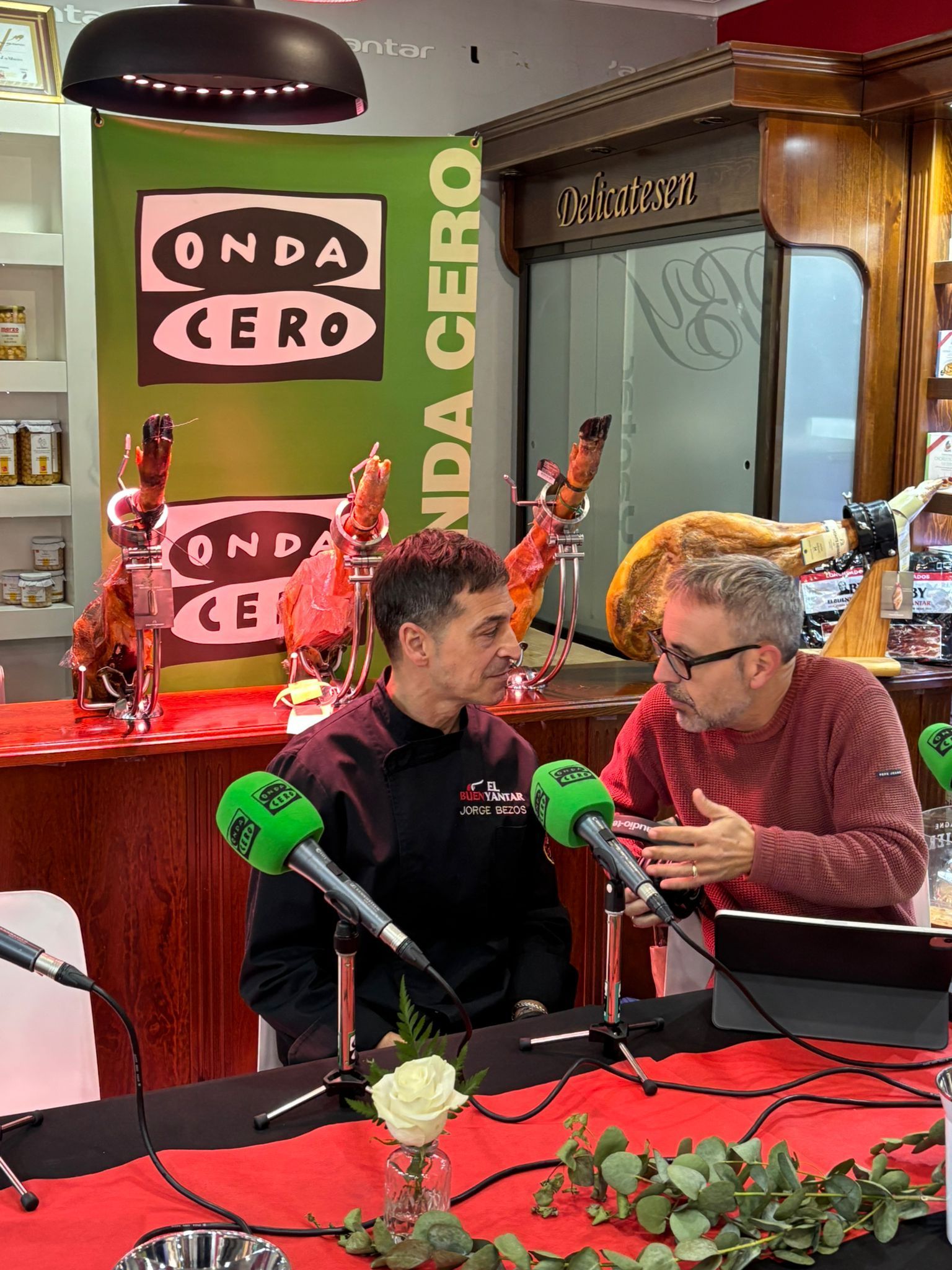 Dos hombres conversan en una mesa de mezclas roja de un estudio de radio Onda Cero, adornada con micrófonos y flores.