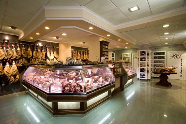 Carnicería con vitrina de cristal para la carne, cortes colgados y estantes refrigerados dentro de una tienda luminosa.