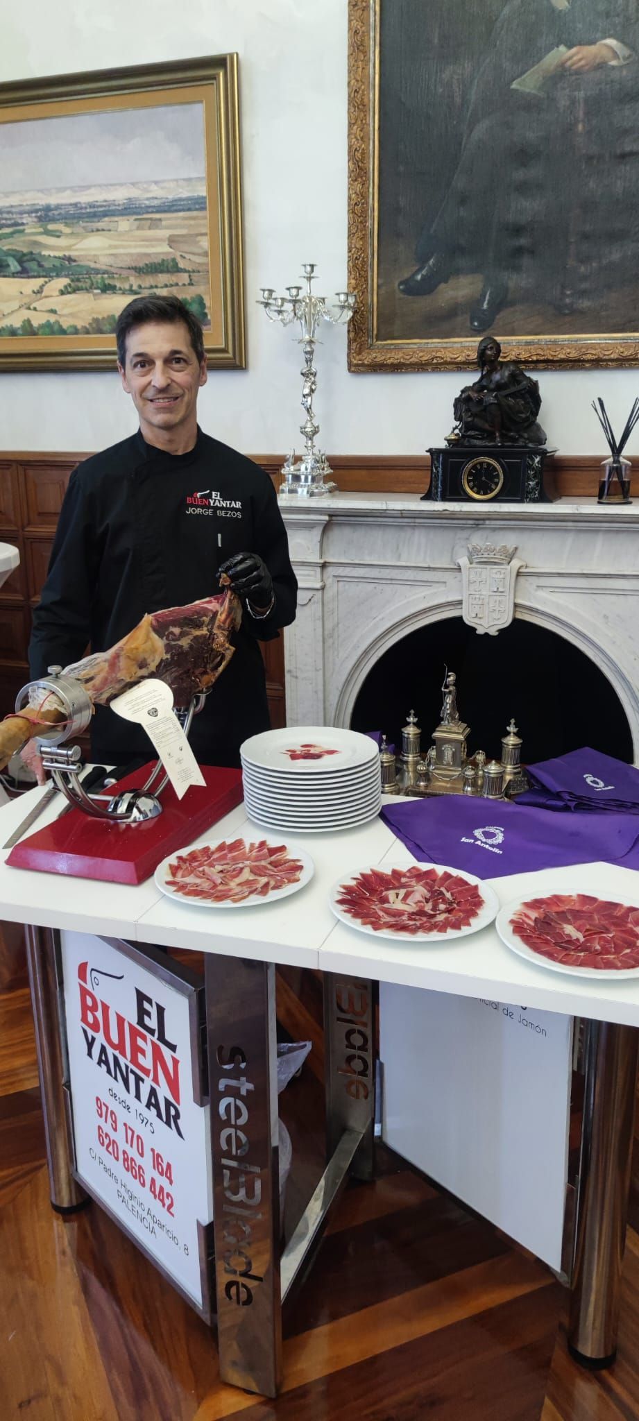 Un hombre presenta pasteles en una mesa con objetos rojos y morados en una habitación ornamentada.