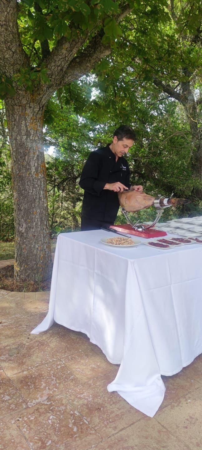 Una persona sirve comida en una mesa blanca al aire libre, bajo un árbol frondoso.