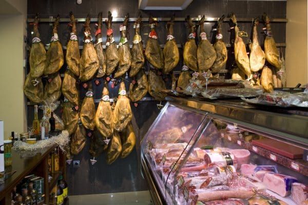 Hileras de jamones curados colgando en una charcutería junto a una vitrina refrigerada para carne.