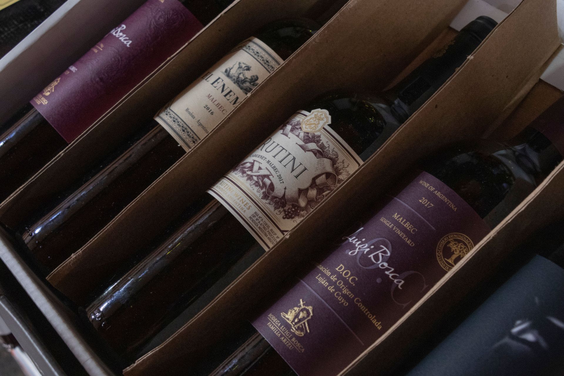 Hilera de botellas de vino en un estante de madera, con etiquetas de color granate y crema visibles.