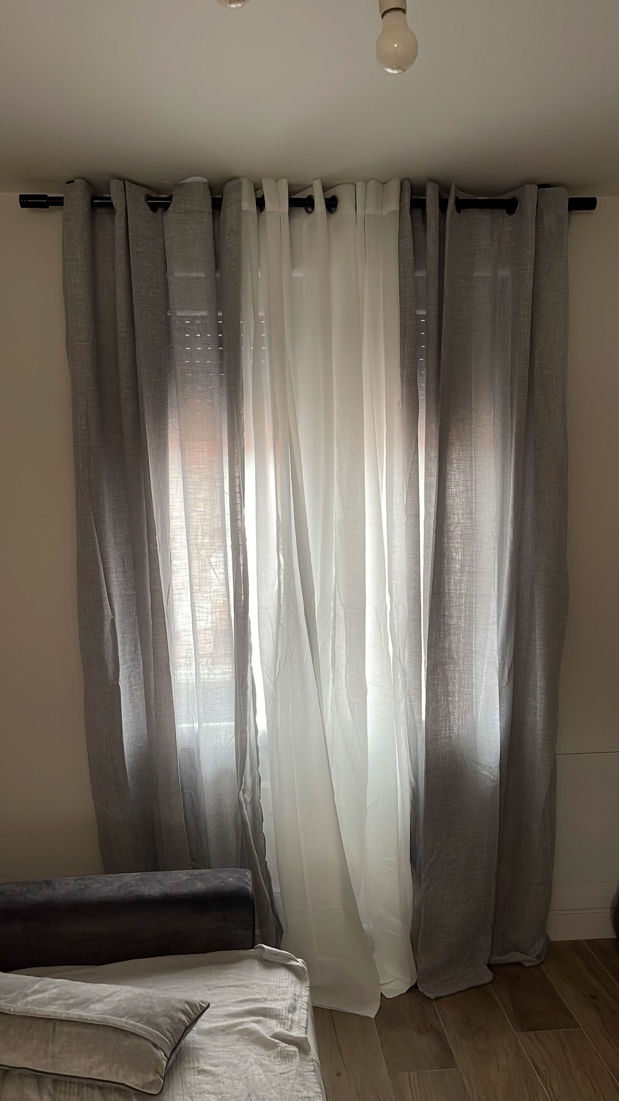 Unas cortinas transparentes de color gris y blanco cuelgan de una barra sobre una ventana, por donde entra la luz.