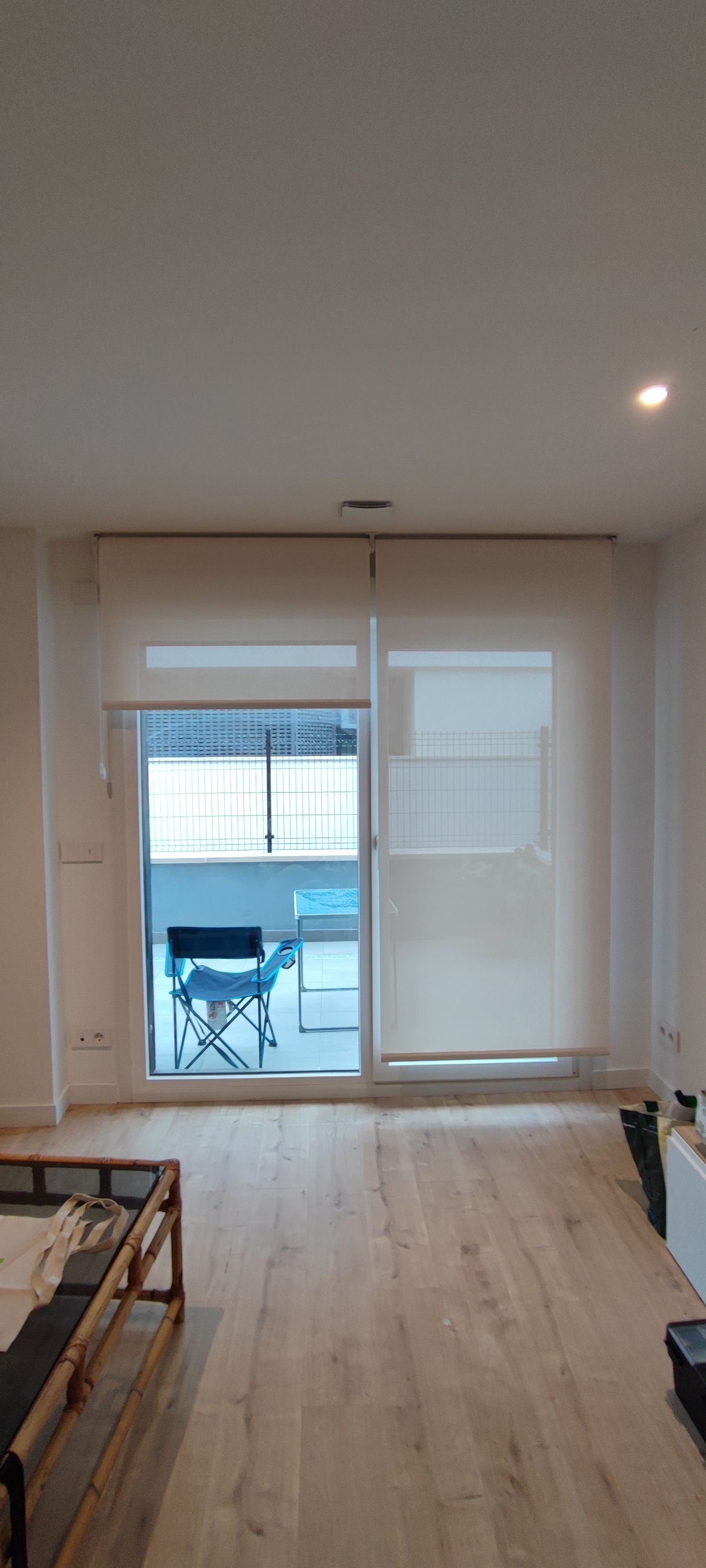 Sala de estar con grandes puertas corredizas de vidrio y persianas blancas. El suelo es de madera clara y se ve un balcón exterior.