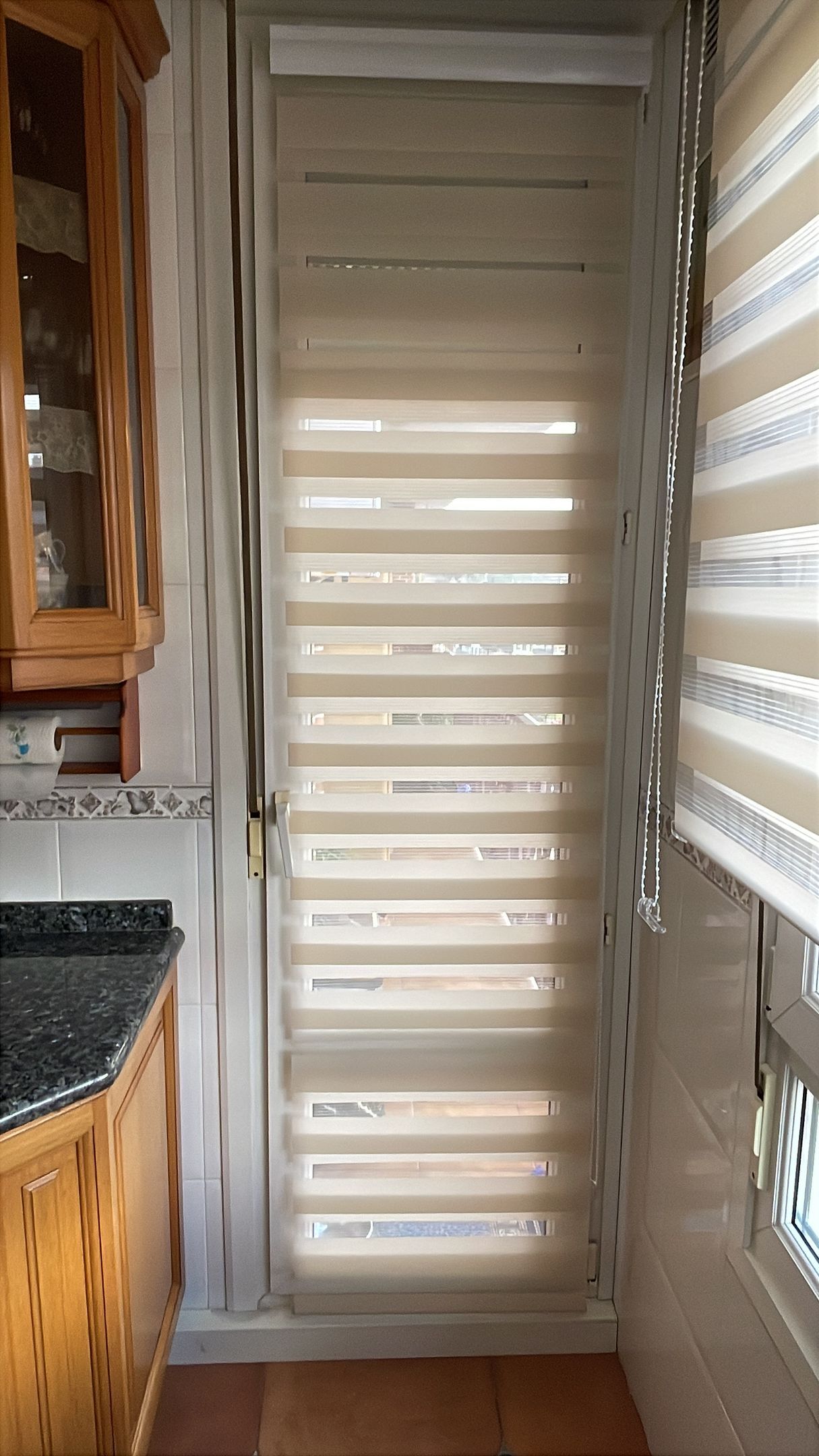 Ventana de la cocina con persianas a rayas, con la luz del sol entrando a raudales por las lamas horizontales. Se ven armarios de madera y una encimera.