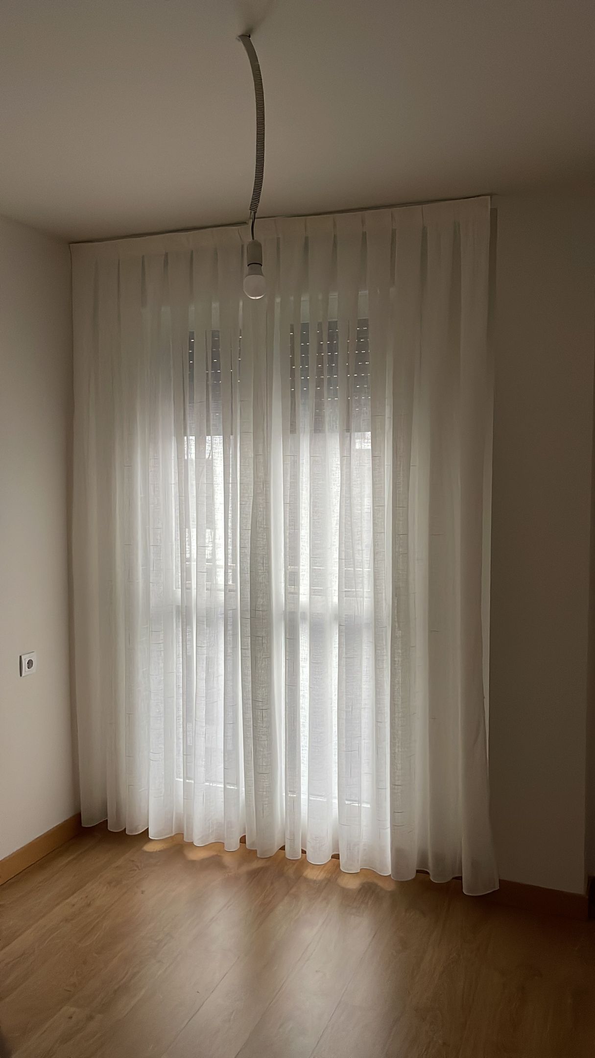 Una ventana con cortinas blancas transparentes en una habitación con suelo de madera. Una lámpara cuelga del techo.