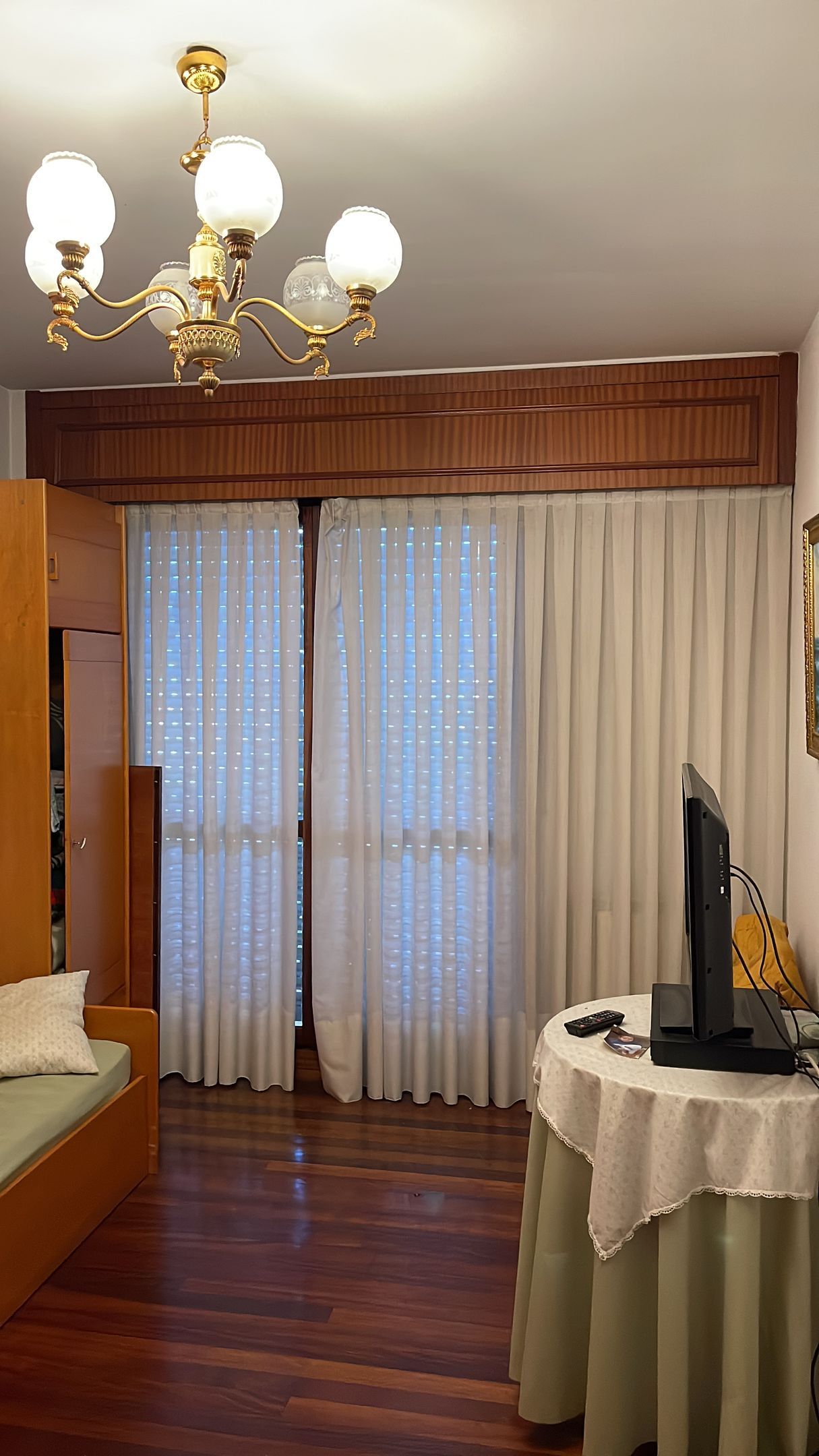 Sala de estar con suelos de madera, cortinas blancas y un pequeño televisor sobre una mesa redonda. Una lámpara de araña cuelga del techo.