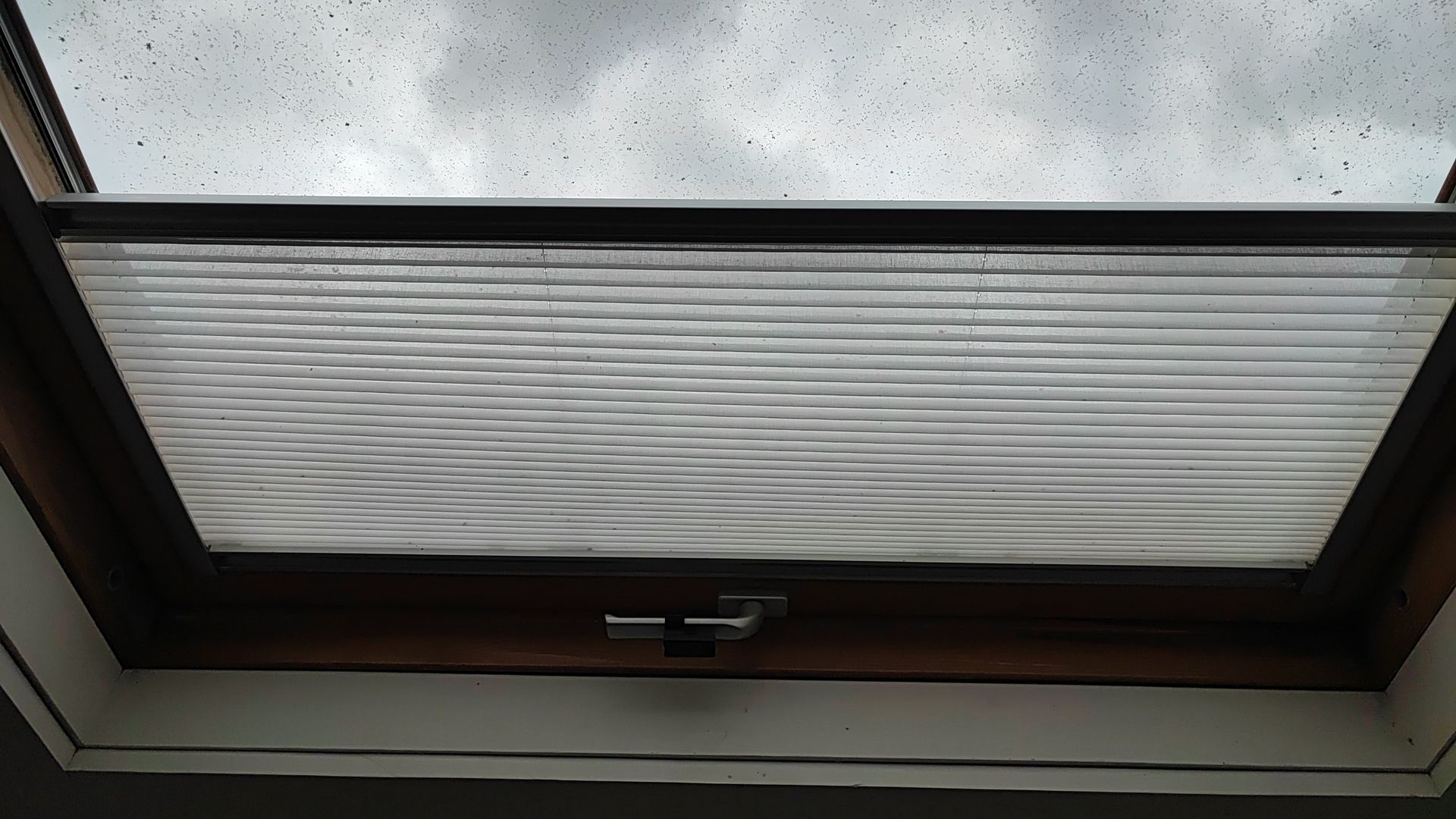 Claraboya con persiana plisada parcialmente extendida. Se ve la lluvia en la ventana. Marco de madera.