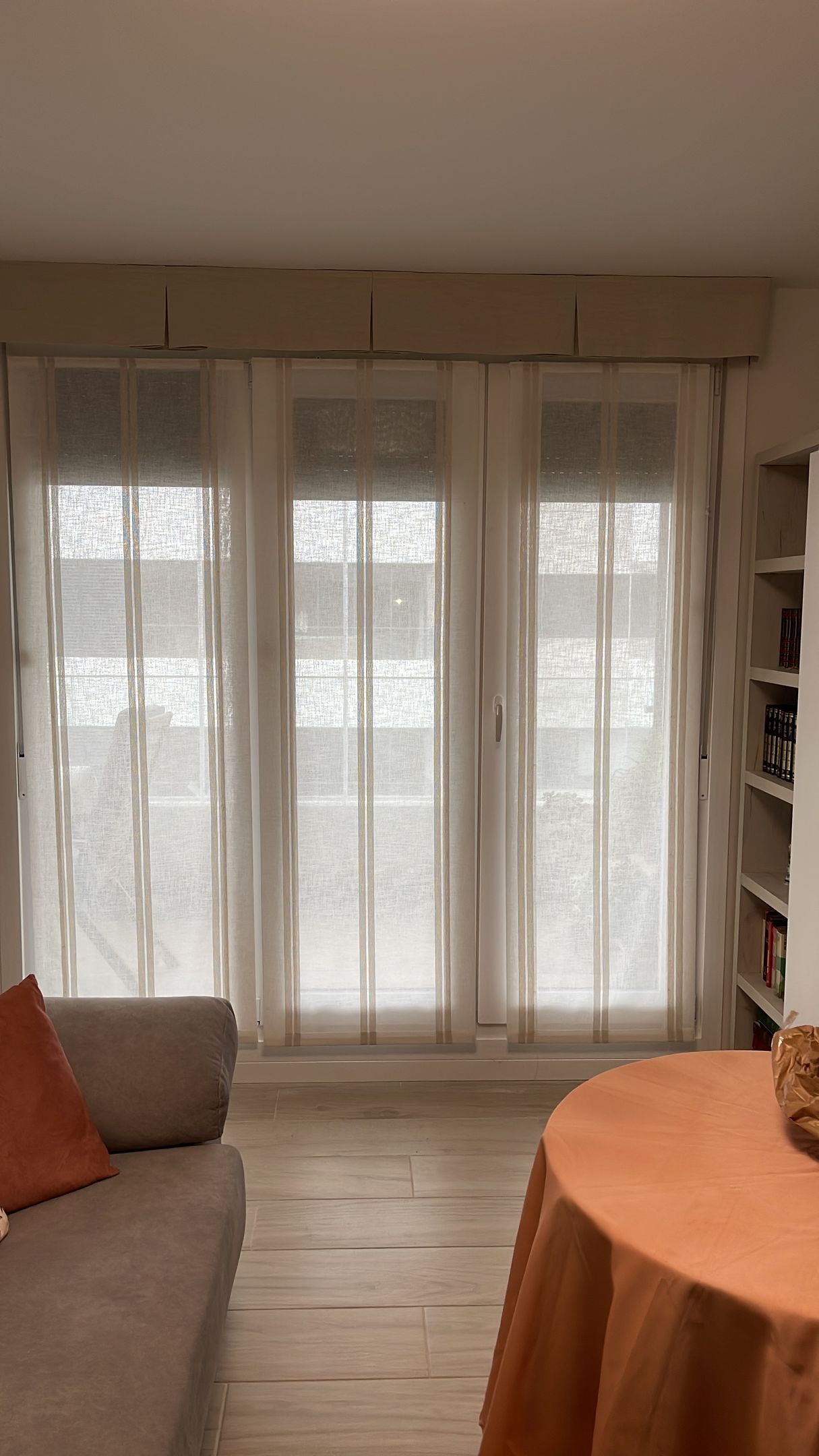 Una sala de estar con tres ventanas cubiertas por cortinas transparentes, un sofá gris y un mantel naranja.