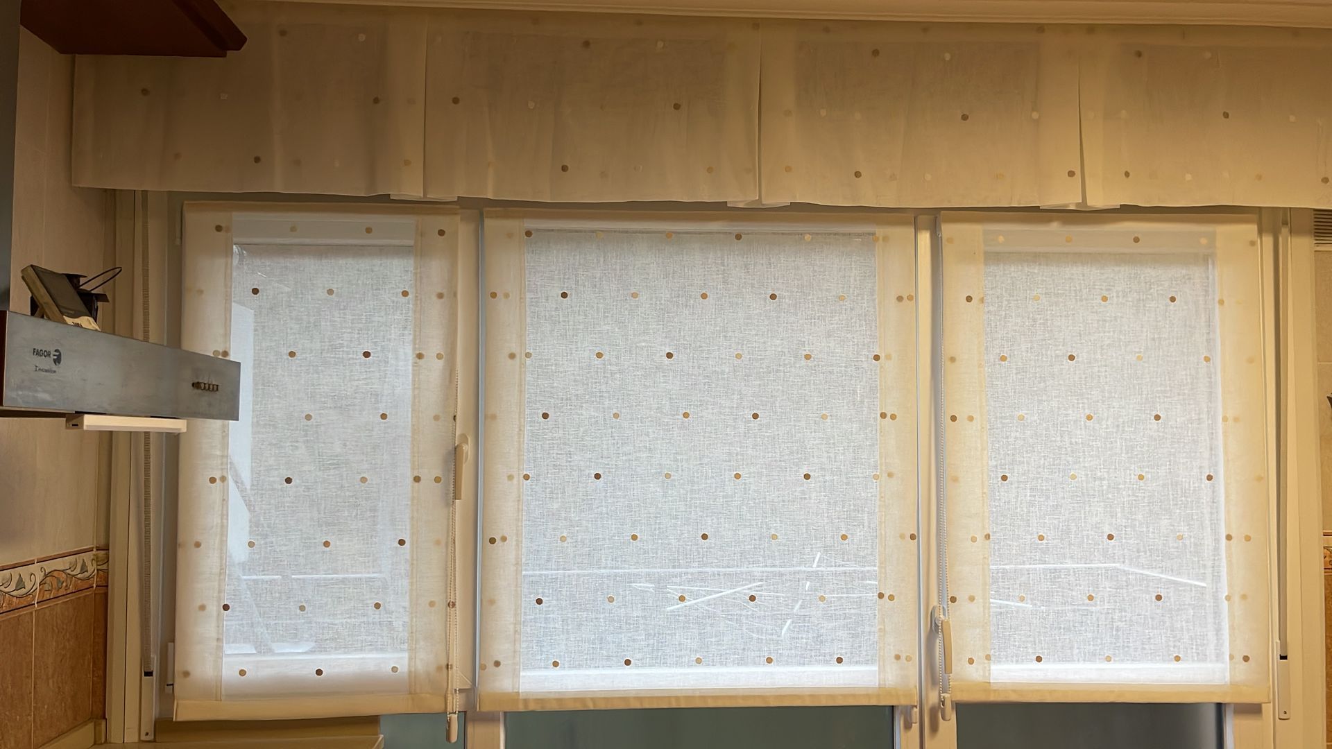 Tres persianas enrollables de color claro en fila, con un faldón a juego encima, en una habitación con ventana.