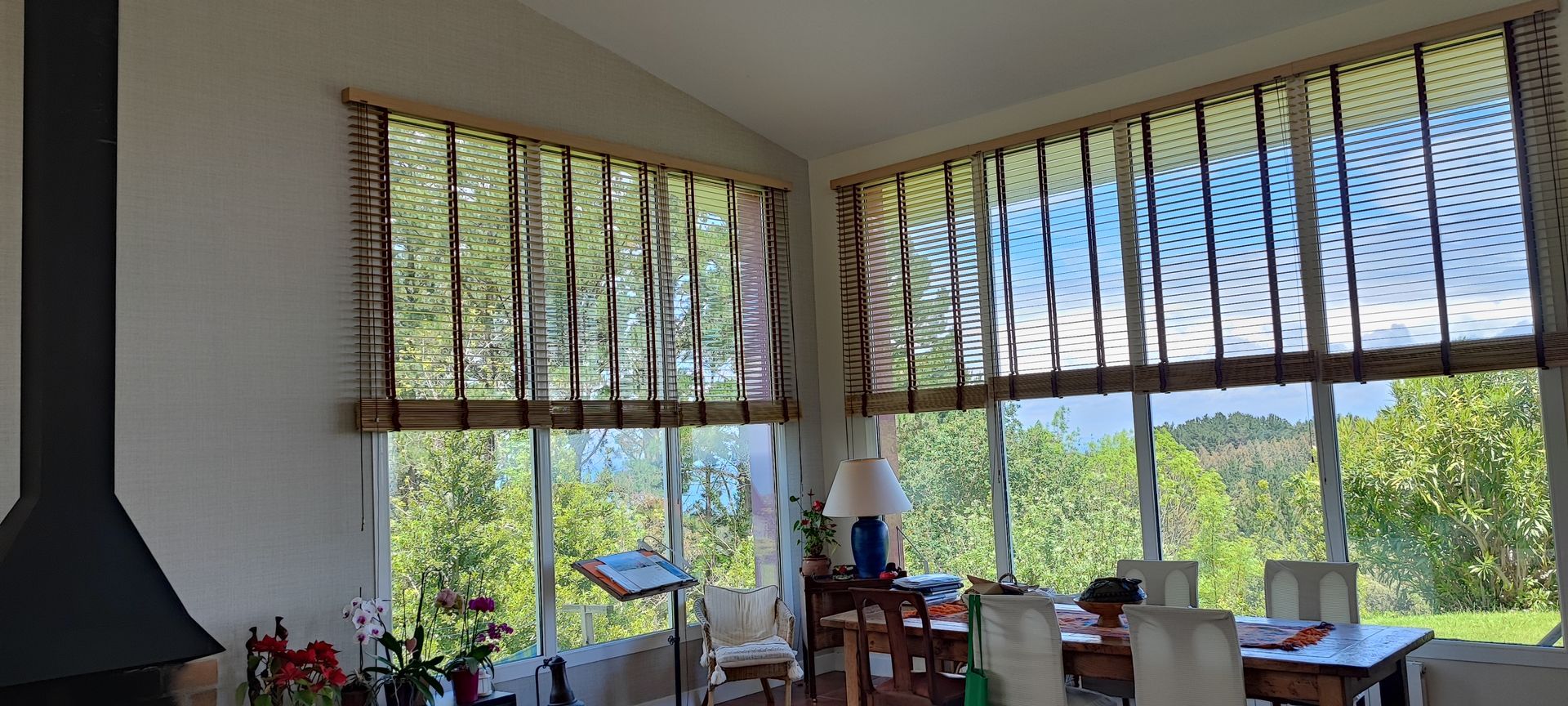 Habitación interior con grandes ventanales, persianas decorativas y vistas a árboles verdes y cielo azul. Se ven una chimenea y muebles en el interior.