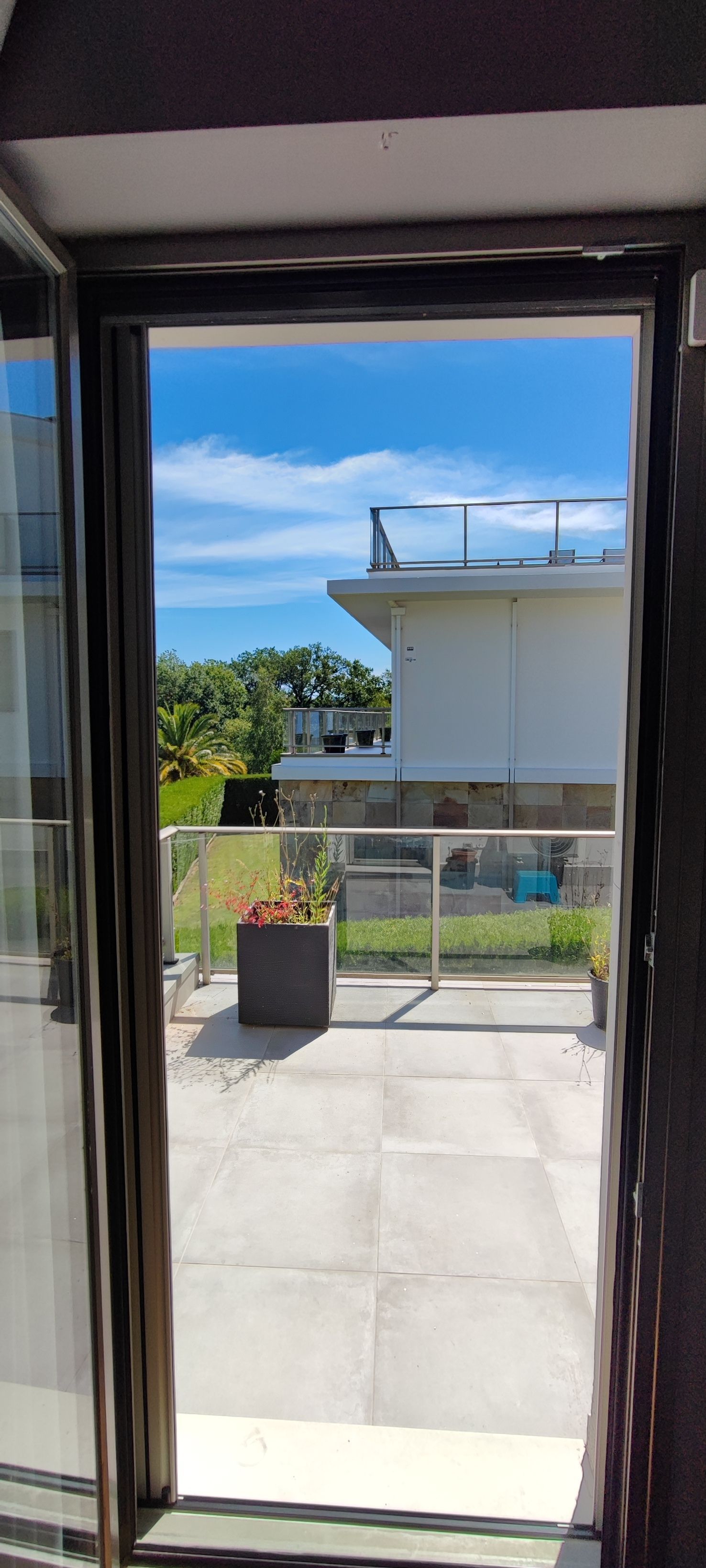 Vista desde una puerta abierta a una terraza con un edificio blanco, cielo azul y un macetero de flores.