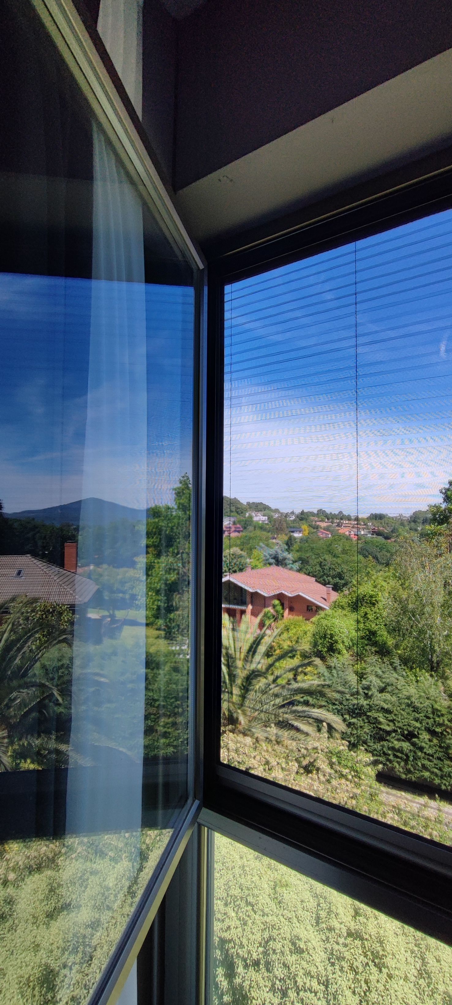 Una ventana abierta con vistas a un barrio con casas de techos rojos y exuberante vegetación bajo un cielo azul.