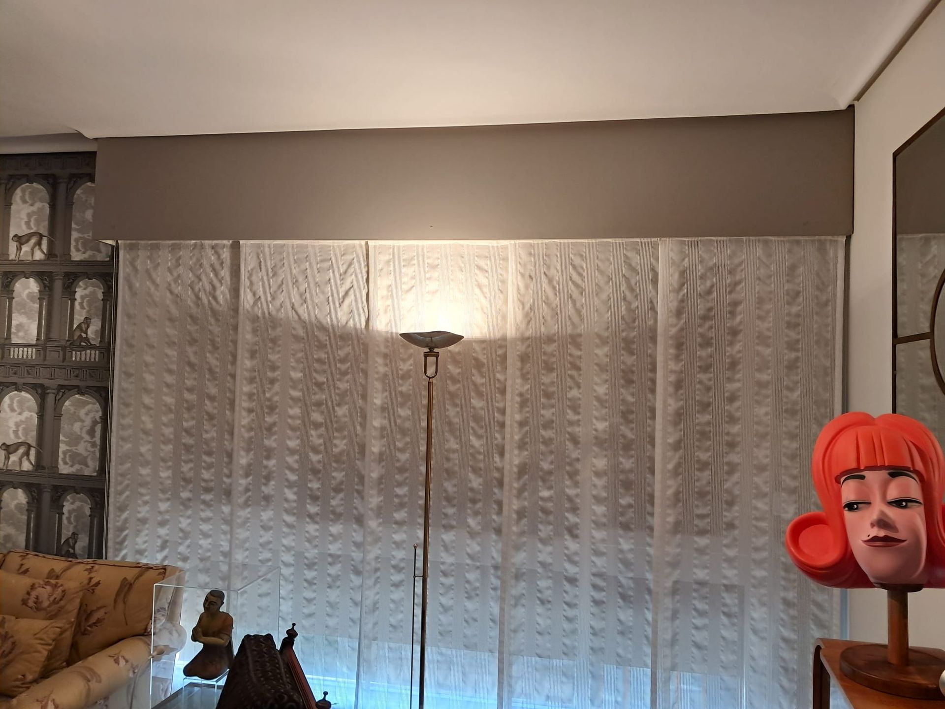 Una habitación con cortinas estampadas, una lámpara de pie y un busto decorativo. Las cortinas tienen un diseño texturizado y un panel marrón oscuro las cubre.