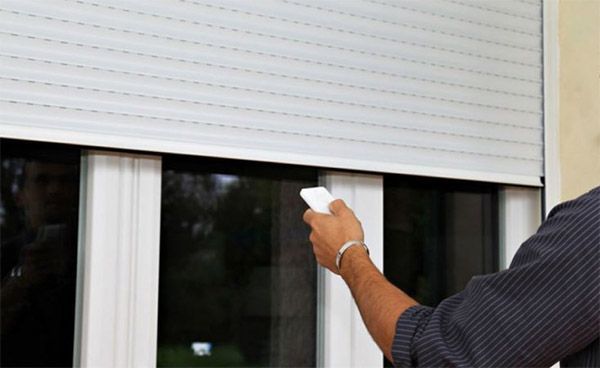 Persona que opera un control remoto blanco, probablemente para subir o bajar una persiana exterior blanca enrollable en una ventana.